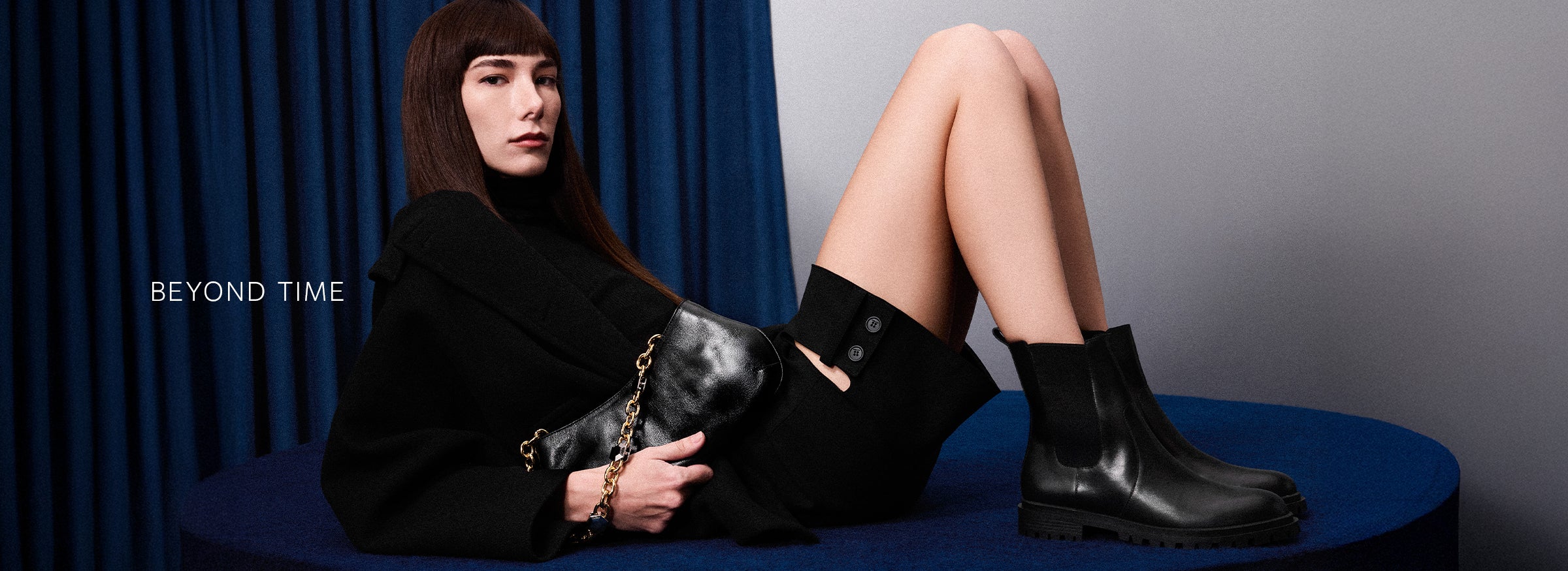 A widescreen image showing a model sitting on a blue surface in front of blue curtains, wearing a black outfit and black Chelsea boots. The text "BEYOND TIME" is on the left.