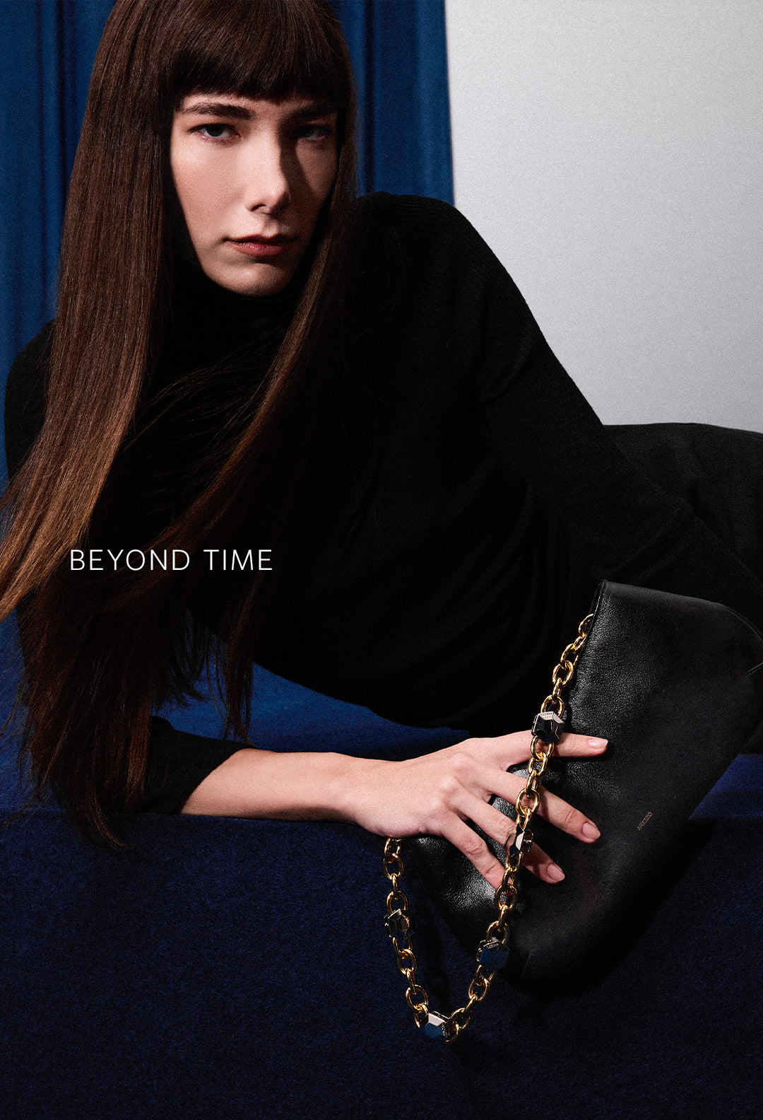 A widescreen image showing a model sitting on a blue surface in front of blue curtains, wearing a black outfit and black Chelsea boots. The text "BEYOND TIME" is on the left.