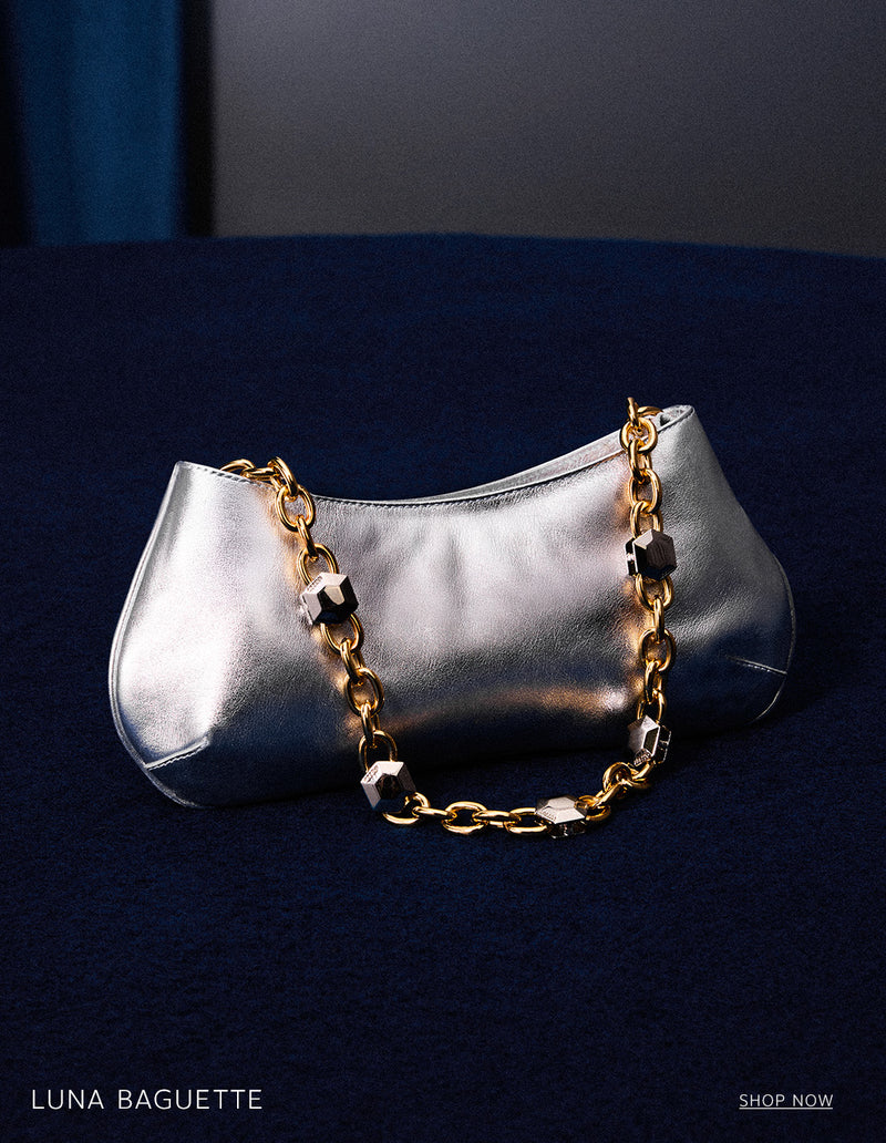 A silver metallic Luna Baguette handbag with a gold chain strap featuring black accents, resting on a dark blue textured surface. The text 