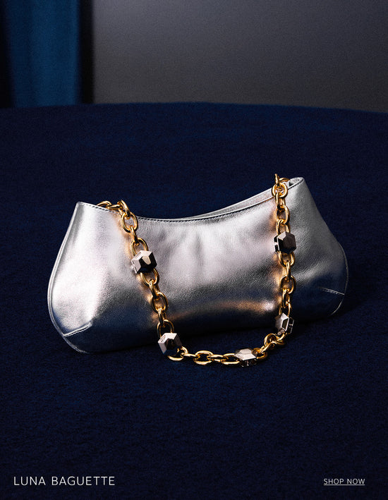 A silver metallic Luna Baguette handbag with a gold chain strap featuring black accents, resting on a dark blue textured surface. The text 