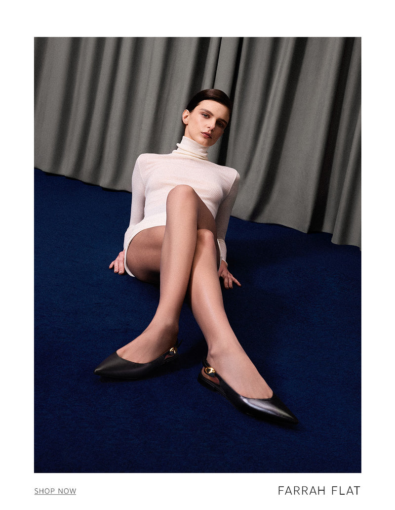 A woman sitting on a blue floor in front of gray curtains, wearing a white long-sleeve top and black Farrah Flat shoes. The text 