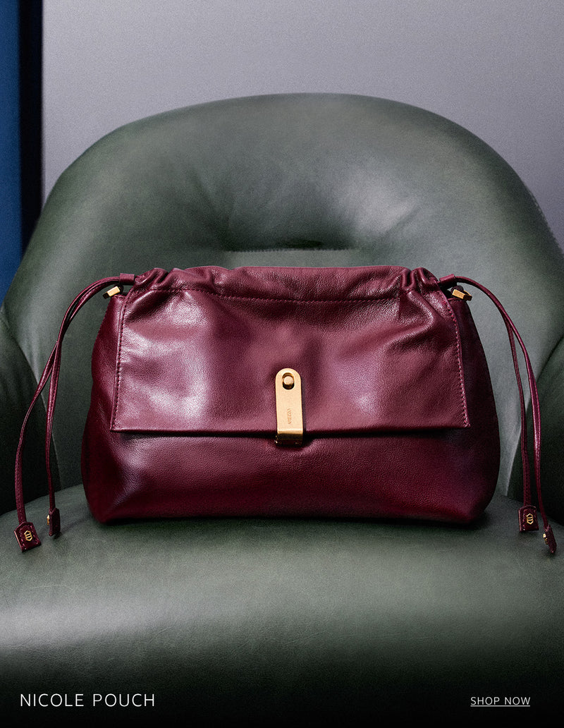 A burgundy leather Nicole Pouch bag with a gold clasp and drawstring details, placed on a green leather armchair. The text 