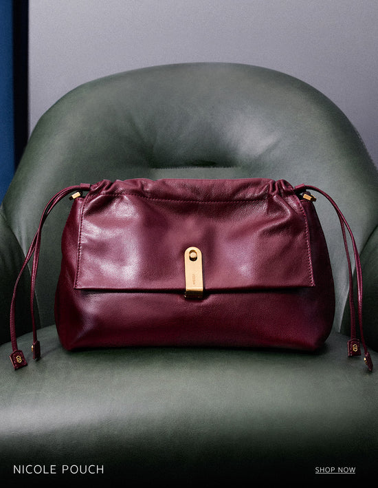 A burgundy leather Nicole Pouch bag with a gold clasp and drawstring details, placed on a green leather armchair. The text 