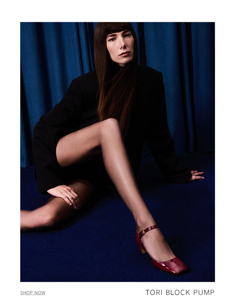 A woman seated on a blue floor in front of blue curtains, wearing a black blazer and sheer black tights, with a visible red patent leather Tori Block Pump on her foot. 