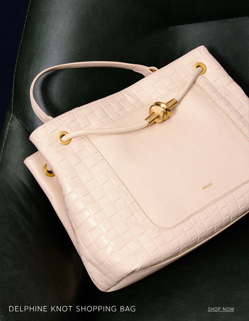 A light pink or beige Delphine Knot Shopping Bag with a woven texture and a gold knot detail on the handle, resting on a dark green leather surface. The text 