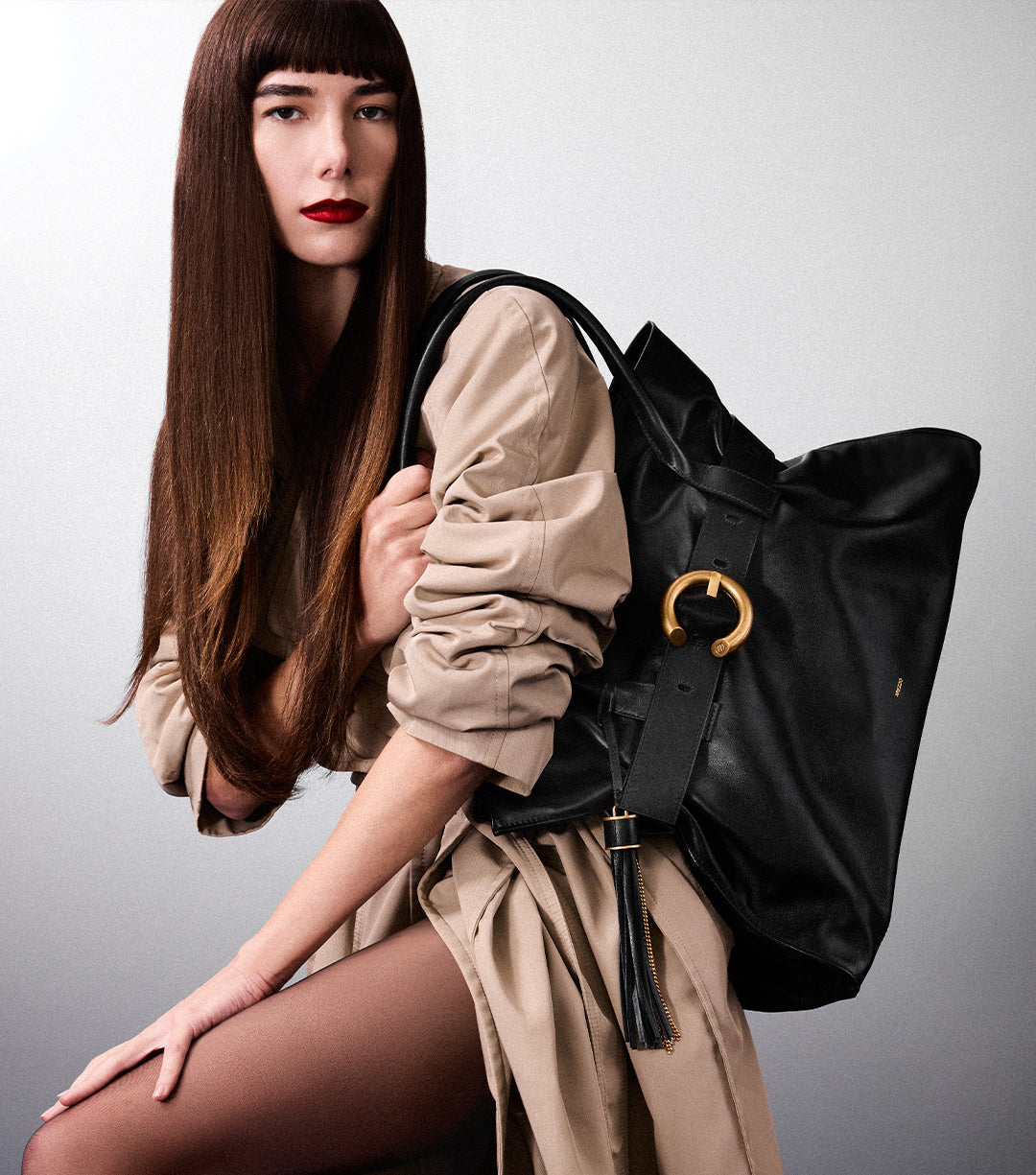 A model with long bangs and red lipstick is shown holding a large black leather bag with a distinctive gold buckle and black tassel.