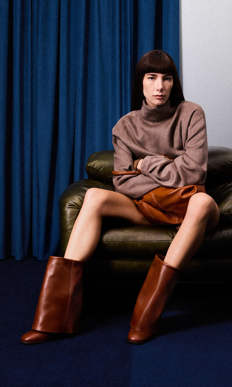 A close-up shot of a model sitting in a green armchair, wearing a brown sweater and a brown leather skirt, holding a matching brown leather handbag with gloves. 
