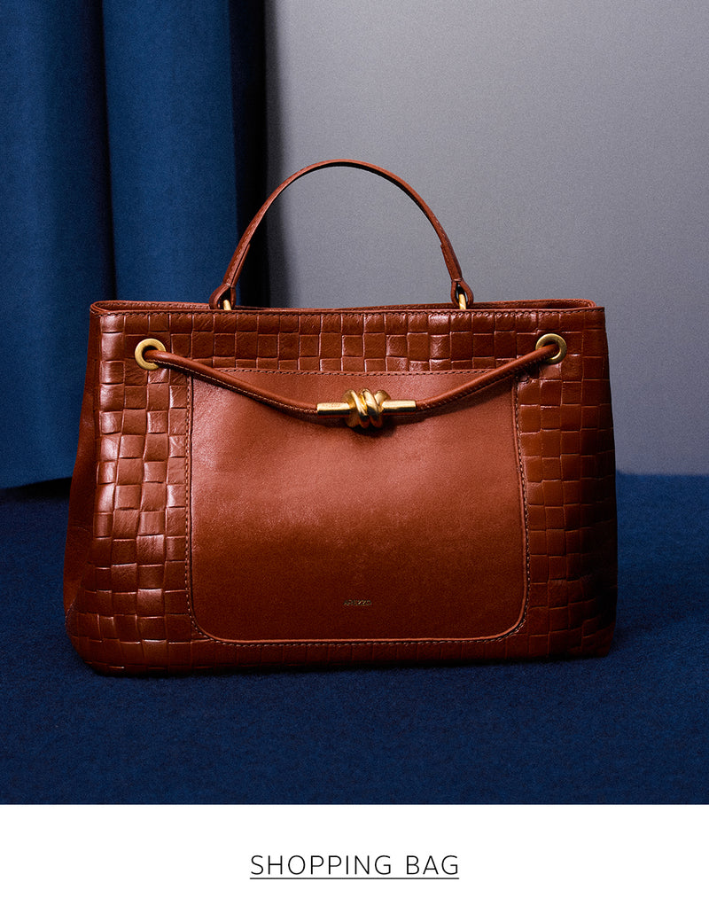 A structured brown shopping bag with a croc-embossed texture and a gold knot detail, resting on a dark blue surface in front of a blue curtain. The text 