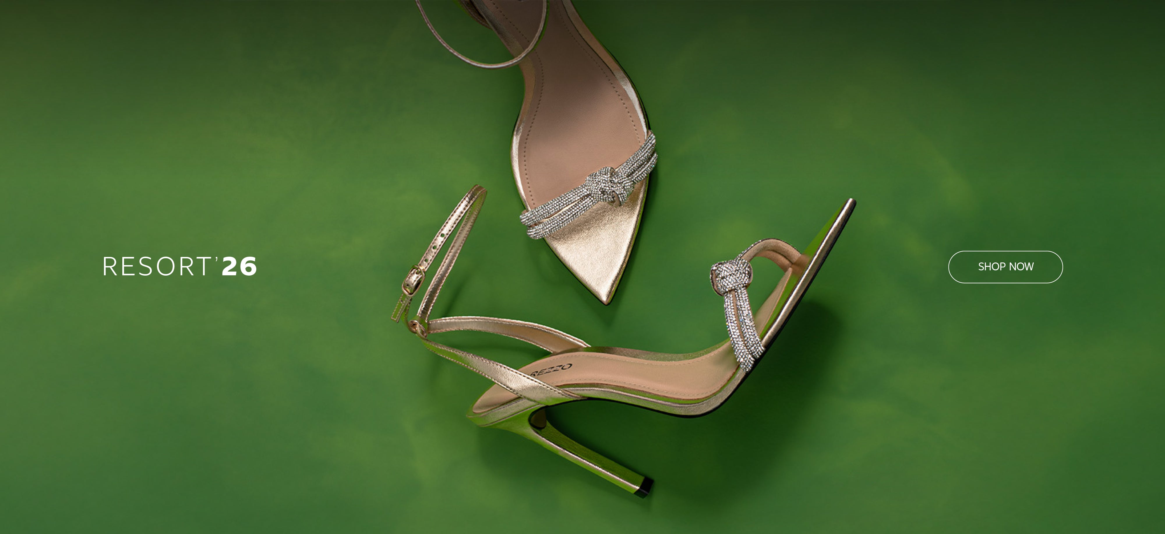 Metallic gold high-heel sandals with crystal-embellished straps displayed on a vibrant green background, promoting the Resort ’26 collection.