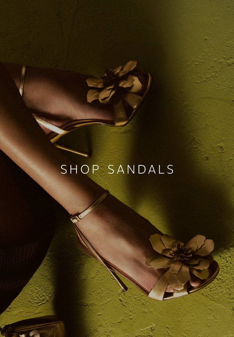 Anne Stiletto Sandal. The style features a high stiletto heel and a rounded toe. It has twisted gold and silver straps over the toes and another diagonal strap across the instep. With a buckle closure and a golden insole with the brand name engraved on it. The style shows the foot. 