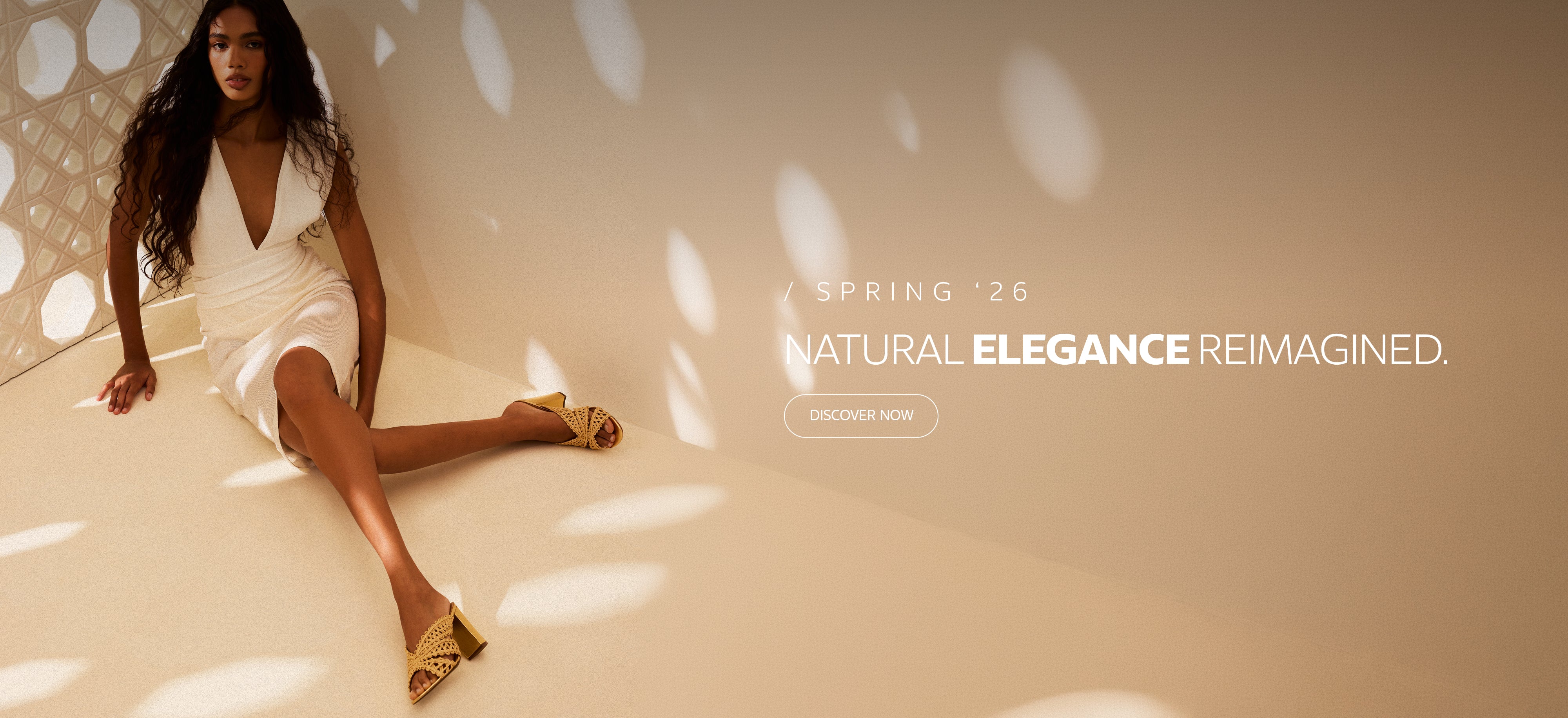 Homepage banner featuring a model wearing a white dress and woven heeled sandals in a warm neutral setting, promoting the Spring ’26 collection ‘Natural Elegance Reimagined’ with a ‘Discover Now’ call to action