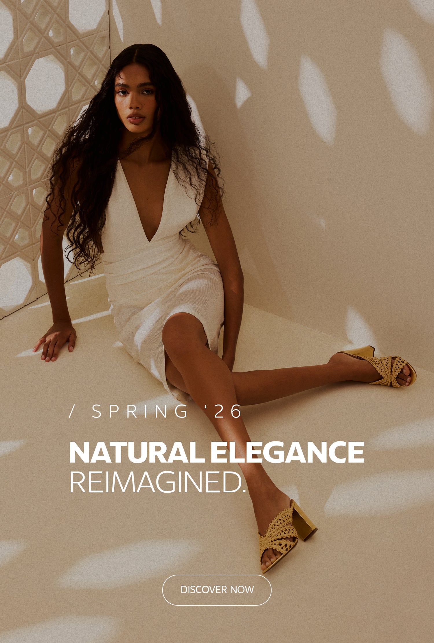 Homepage banner featuring a model wearing a white dress and woven heeled sandals in a warm neutral setting, promoting the Spring ’26 collection ‘Natural Elegance Reimagined’ with a ‘Discover Now’ call to action