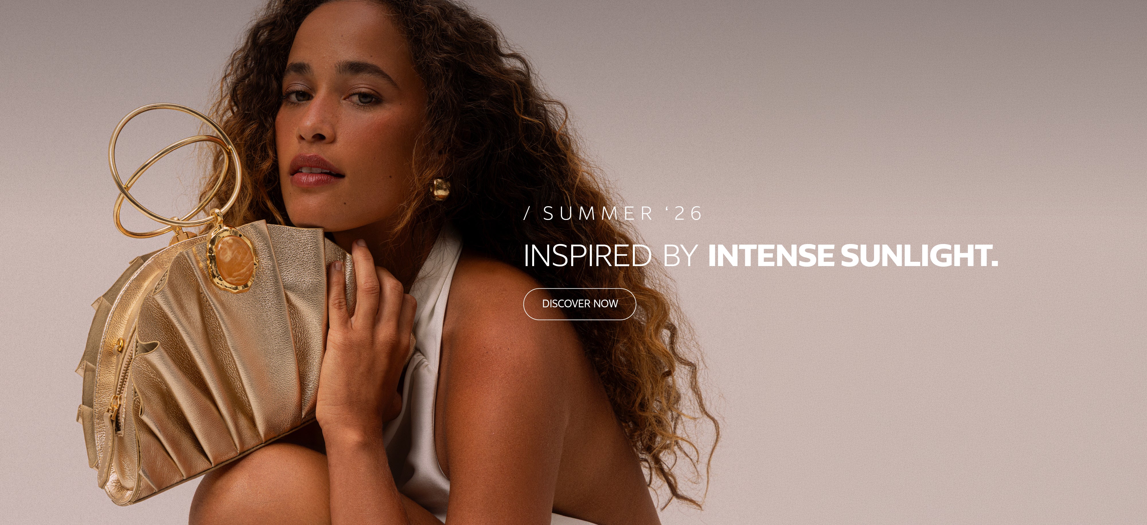 Homepage banner featuring a model holding a gold handbag, promoting the Summer ’26 collection ‘Inspired by Intense Sunlight’ with a ‘Discover Now’ call to action.