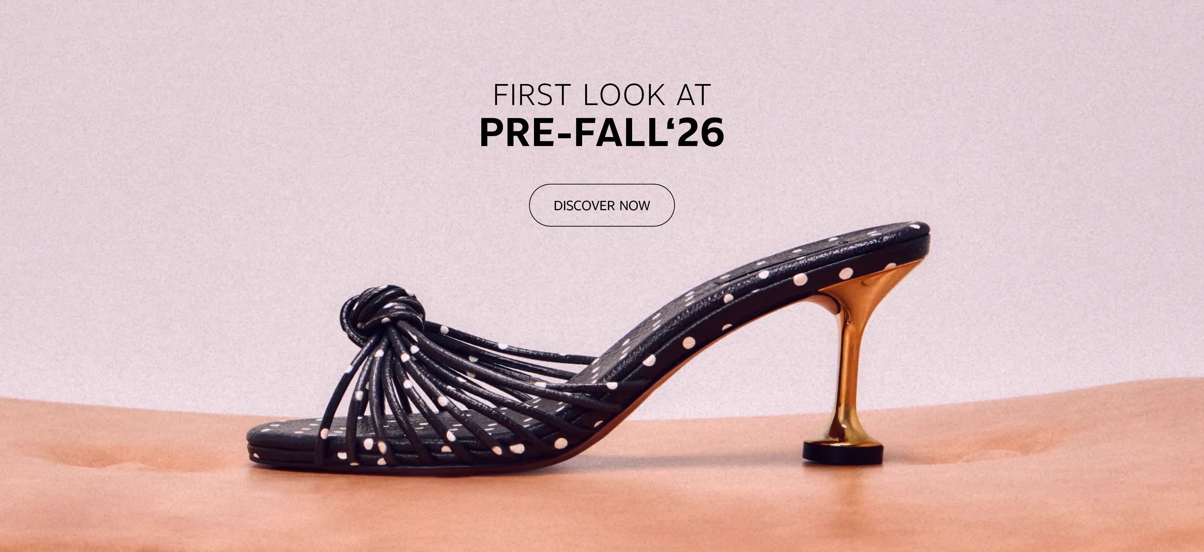 Black polka-dot heeled sandal with knotted strap detailing and a metallic gold heel, displayed on a soft peach pedestal against a neutral backdrop. Above the shoe, the text reads "First Look at Pre-Fall '26" with a "Discover Now" call-to-action button.