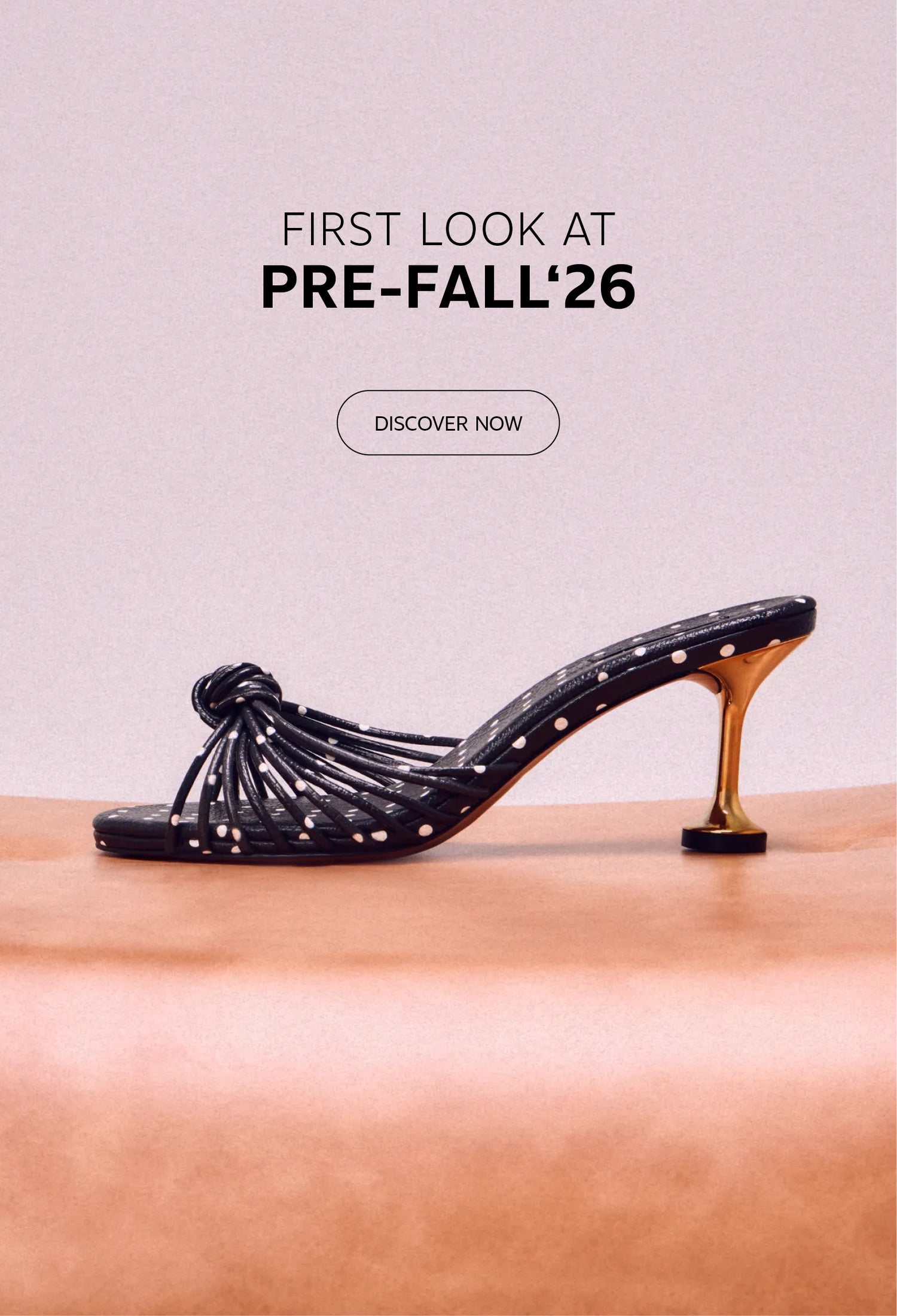 Black polka-dot heeled sandal with knotted strap detailing and a metallic gold heel, displayed on a soft peach pedestal against a neutral backdrop. Above the shoe, the text reads "First Look at Pre-Fall '26" with a "Discover Now" call-to-action button.