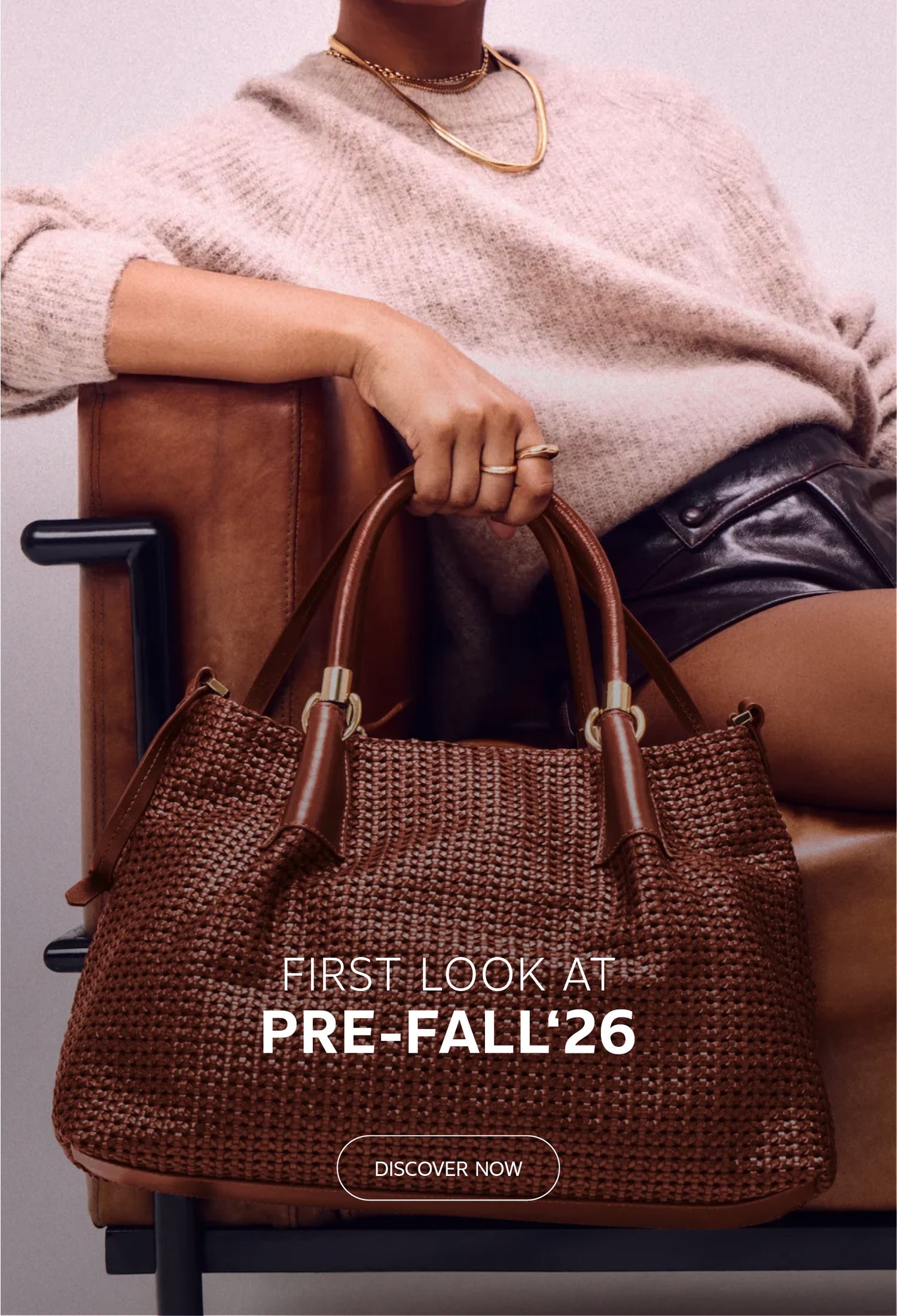 A woman seated on a brown leather armchair, wearing a beige sweater and black leather shorts, holding a large brown woven leather handbag. Prominent white text overlay reads 'FIRST LOOK AT PRE-FALL '26' with a 'DISCOVER NOW' button below. Fashion advertisement style.