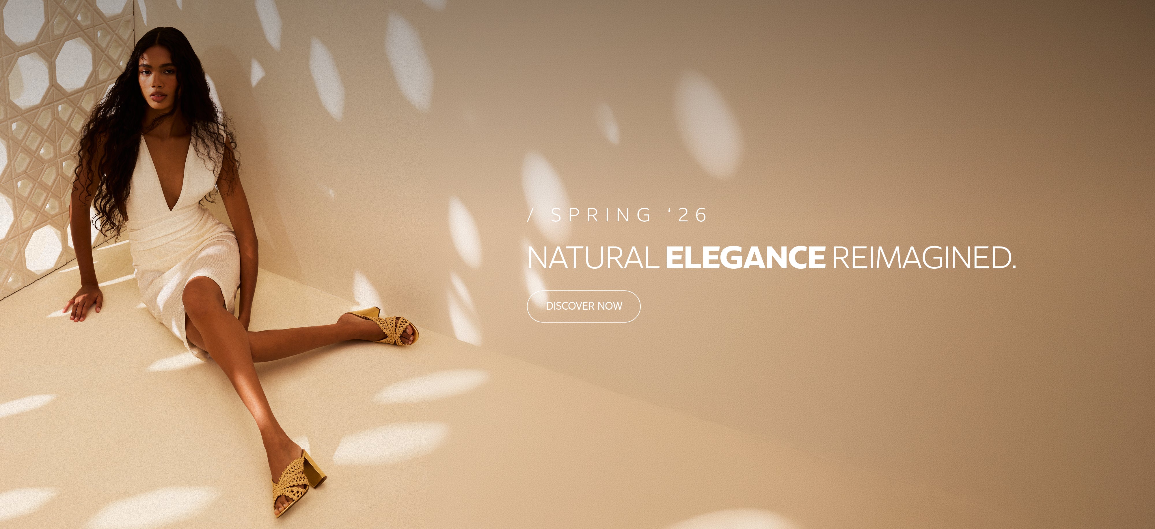 Homepage banner featuring a model wearing a white dress and woven heeled sandals in a warm neutral setting, promoting the Spring ’26 collection ‘Natural Elegance Reimagined’ with a ‘Discover Now’ call to action