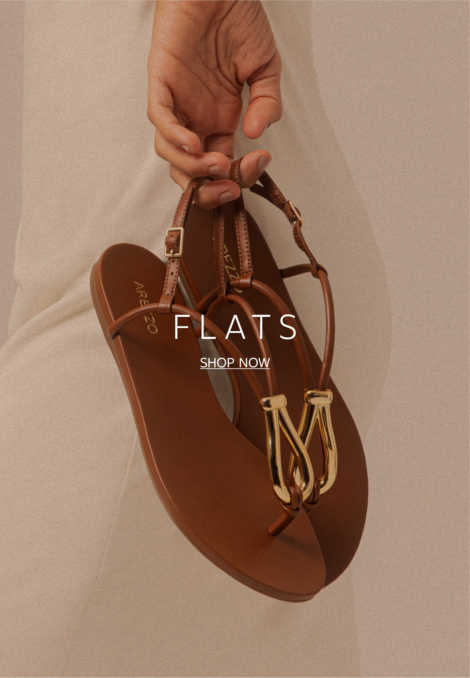 A hand holding a pair of brown leather flat thong sandals by the straps. The sandals feature a distinct, organic-shaped gold metallic buckle detail on the front.