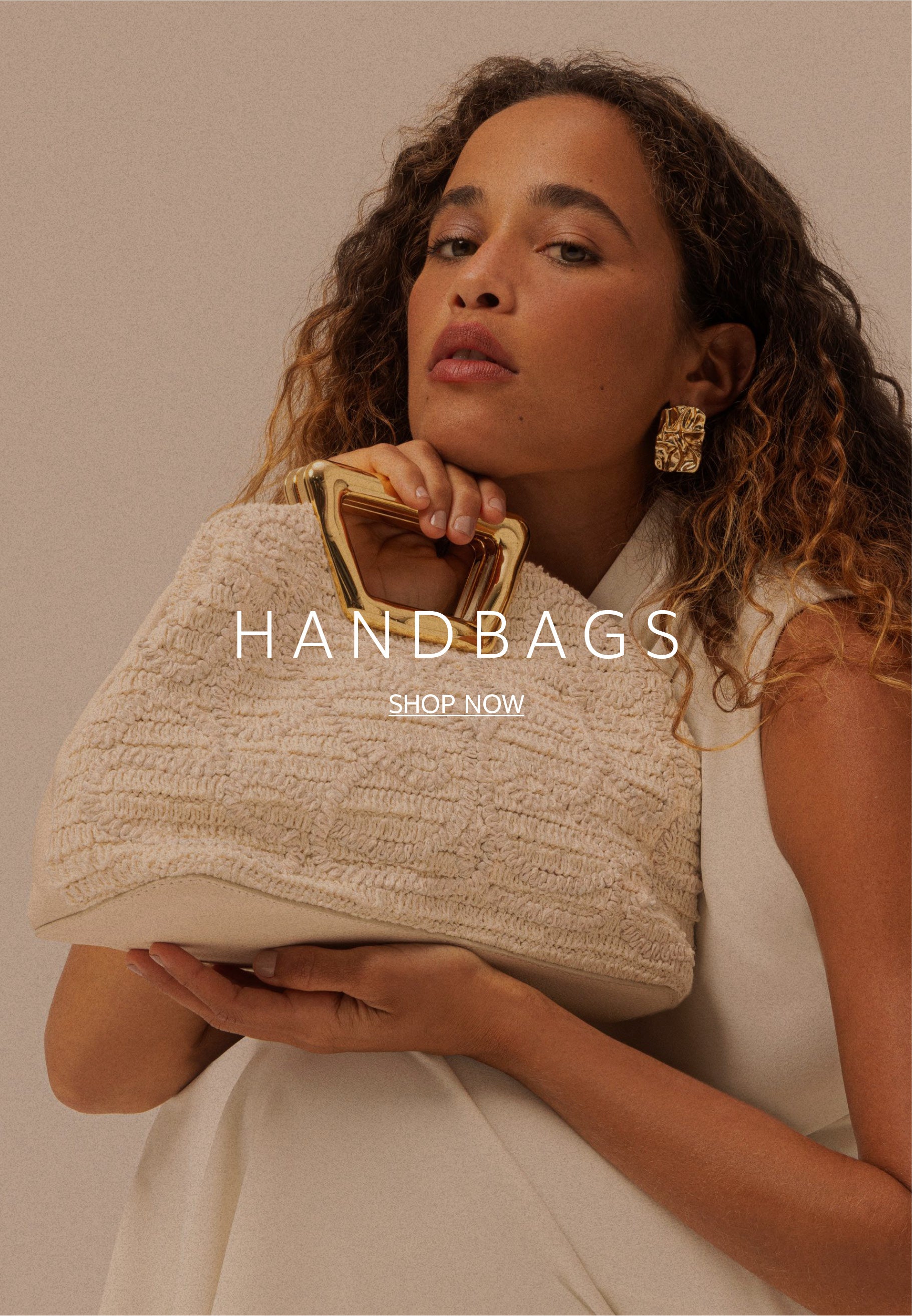 A woman with curly hair posing while holding a textured, off-white fabric clutch handbag featuring a geometric, sculptural gold metal top handle.